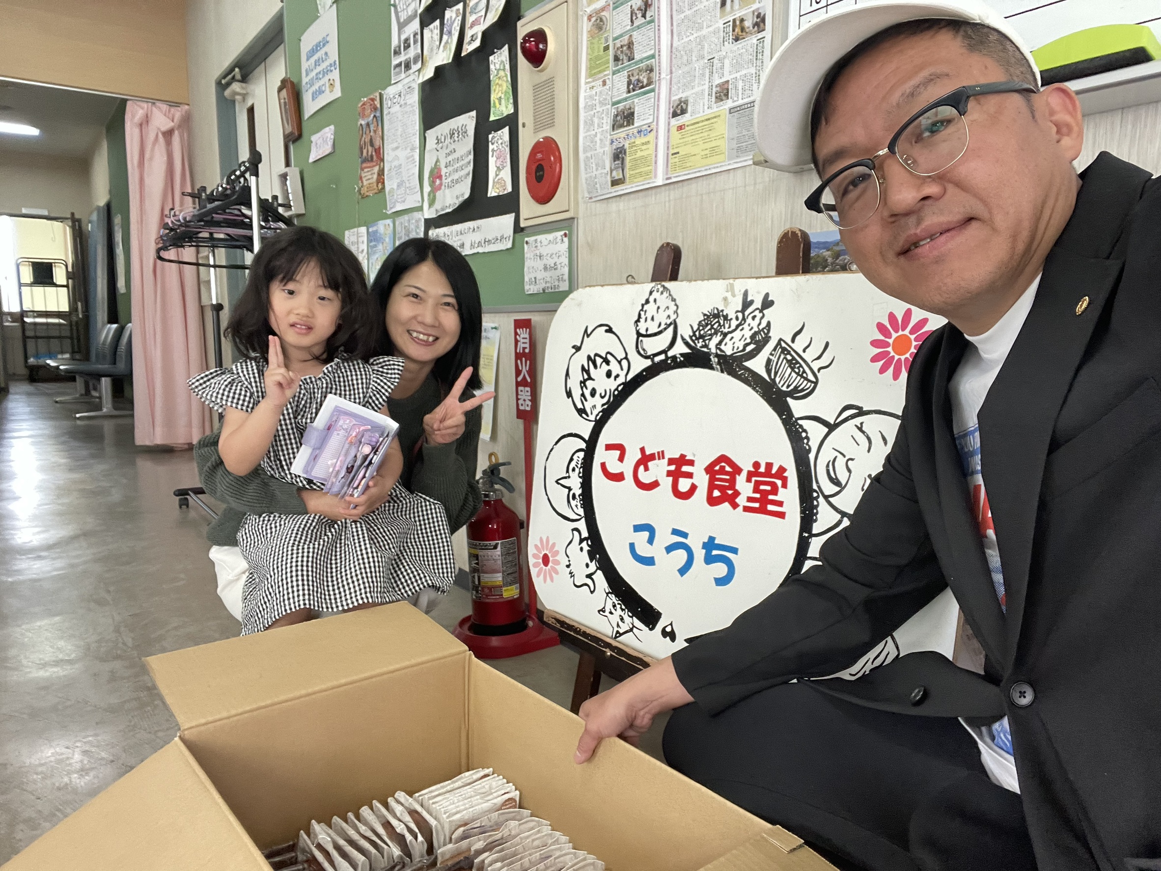 A man in a suit and white cap takes a selfie with a smiling woman and young girl in a community room beside a large sign with Japanese text, near an open cardboard box of packaged items on a table.