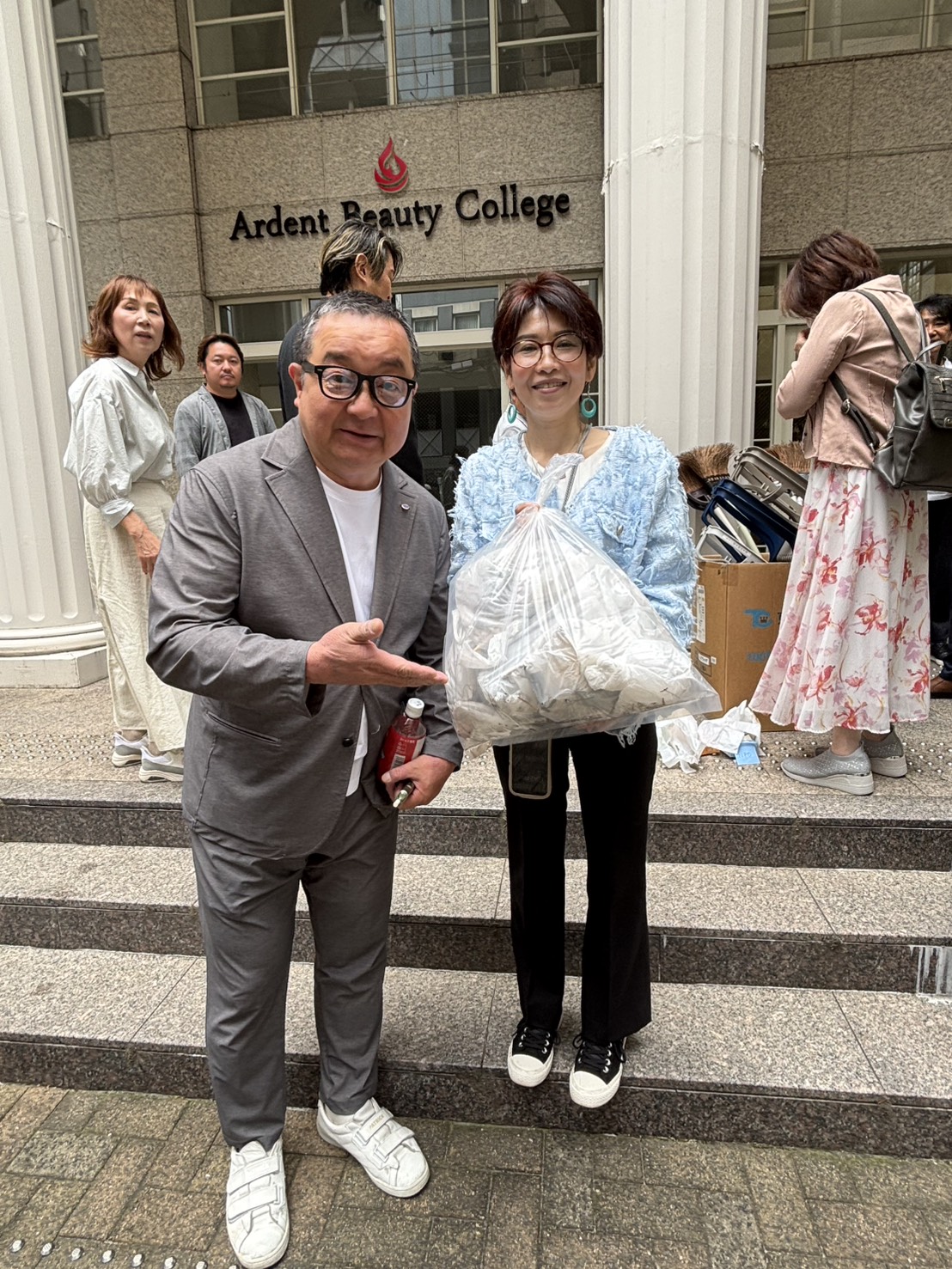 Two people stand on steps outside Ardent Beauty College; a man in a gray suit points to a large plastic bag held by a smiling woman in a light blue jacket, as others donate in the background.