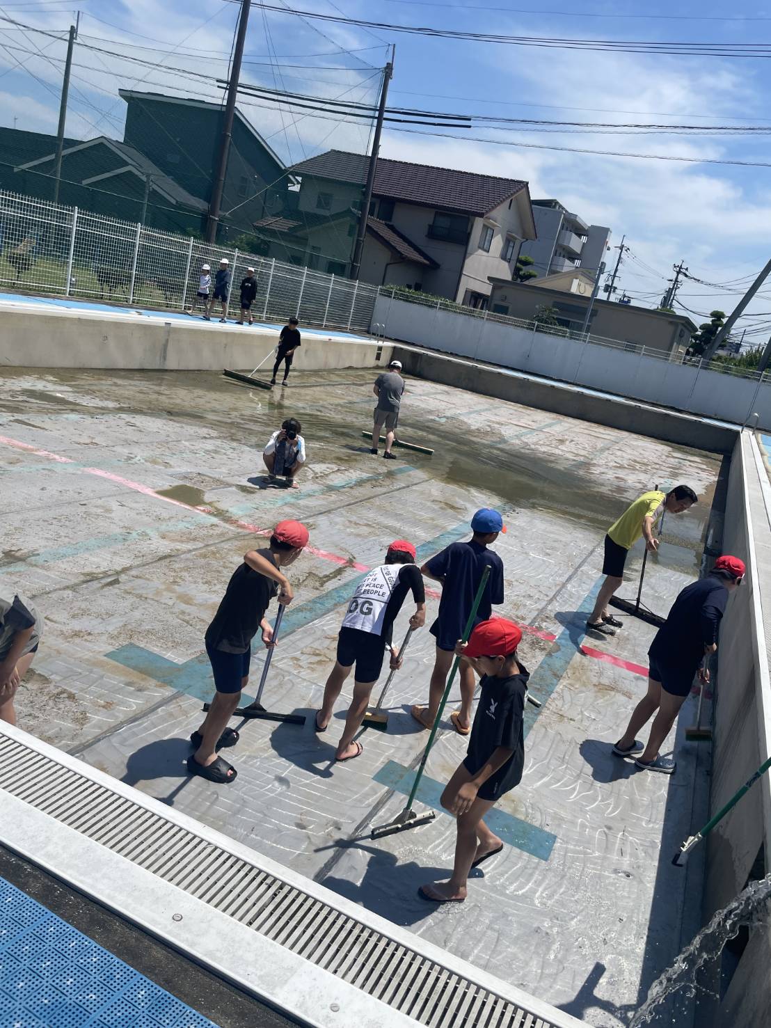Children wearing red caps sweeping a wet concrete pool deck with long-handled brooms under a sunny sky.