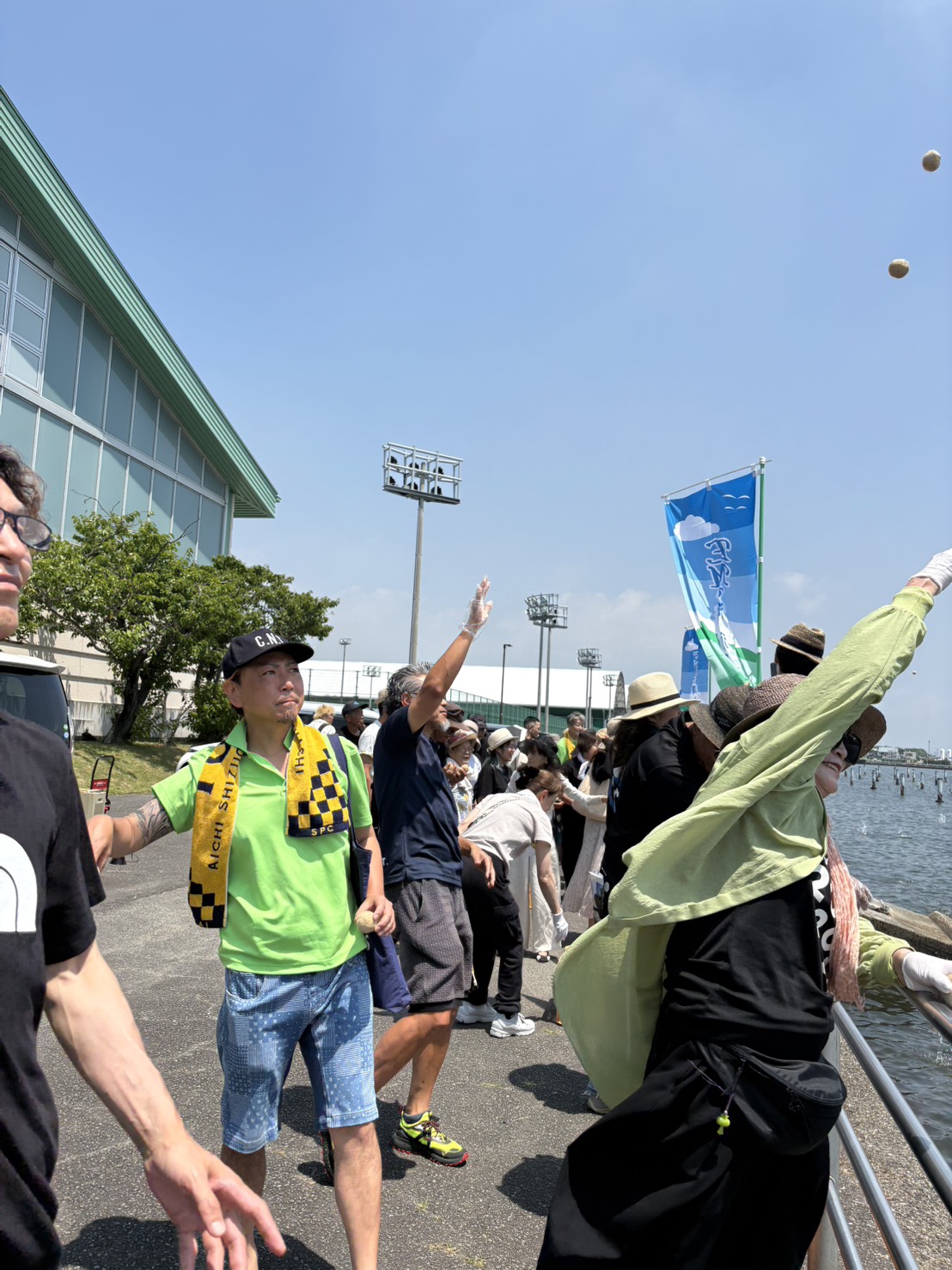 People line a waterfront railing during a sunny outdoor event; some reach toward the water, banners on poles visible in the background.