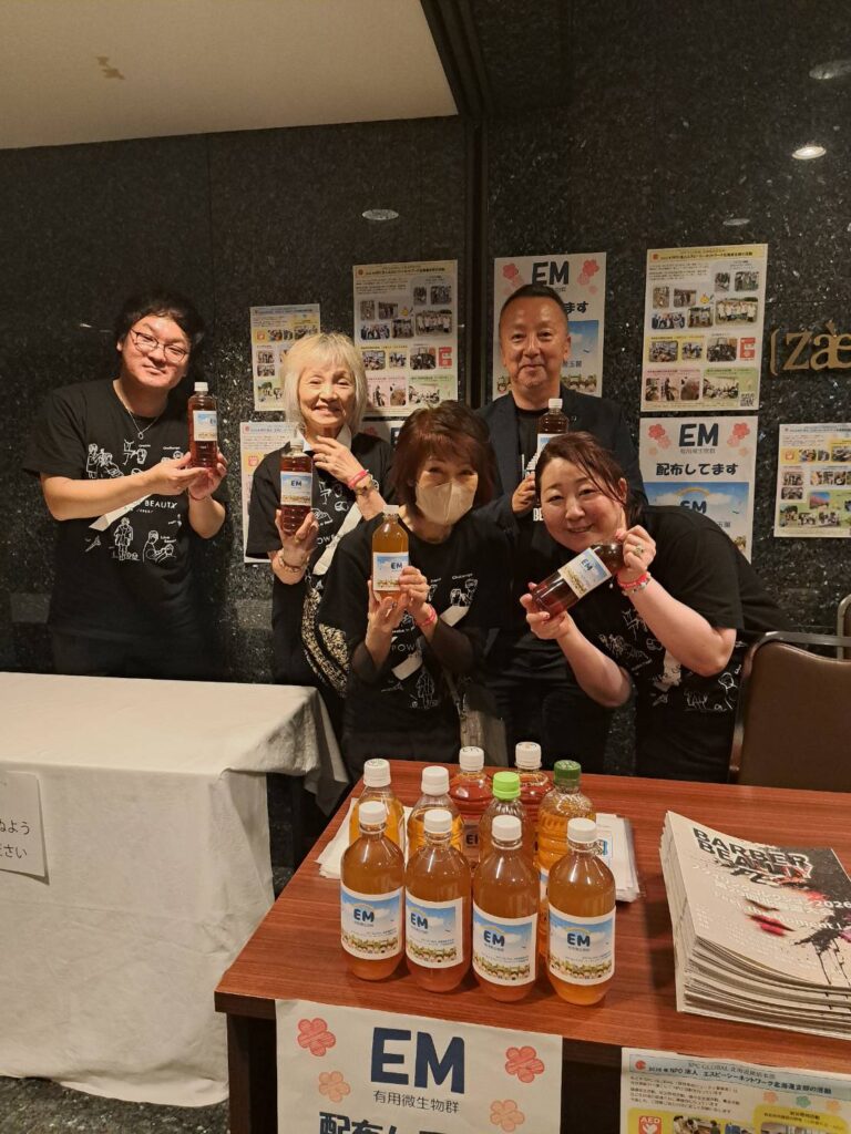 Five people in black shirts stand behind a table, each holding a bottle of EM drink for a booth promotional event with posters on a dark wall behind them.