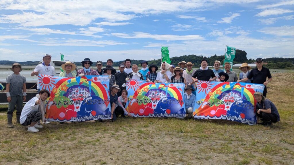Group of about 20 people posing outdoors on a sunny day, holding colorful banners for a community event with a river and hills in the background.