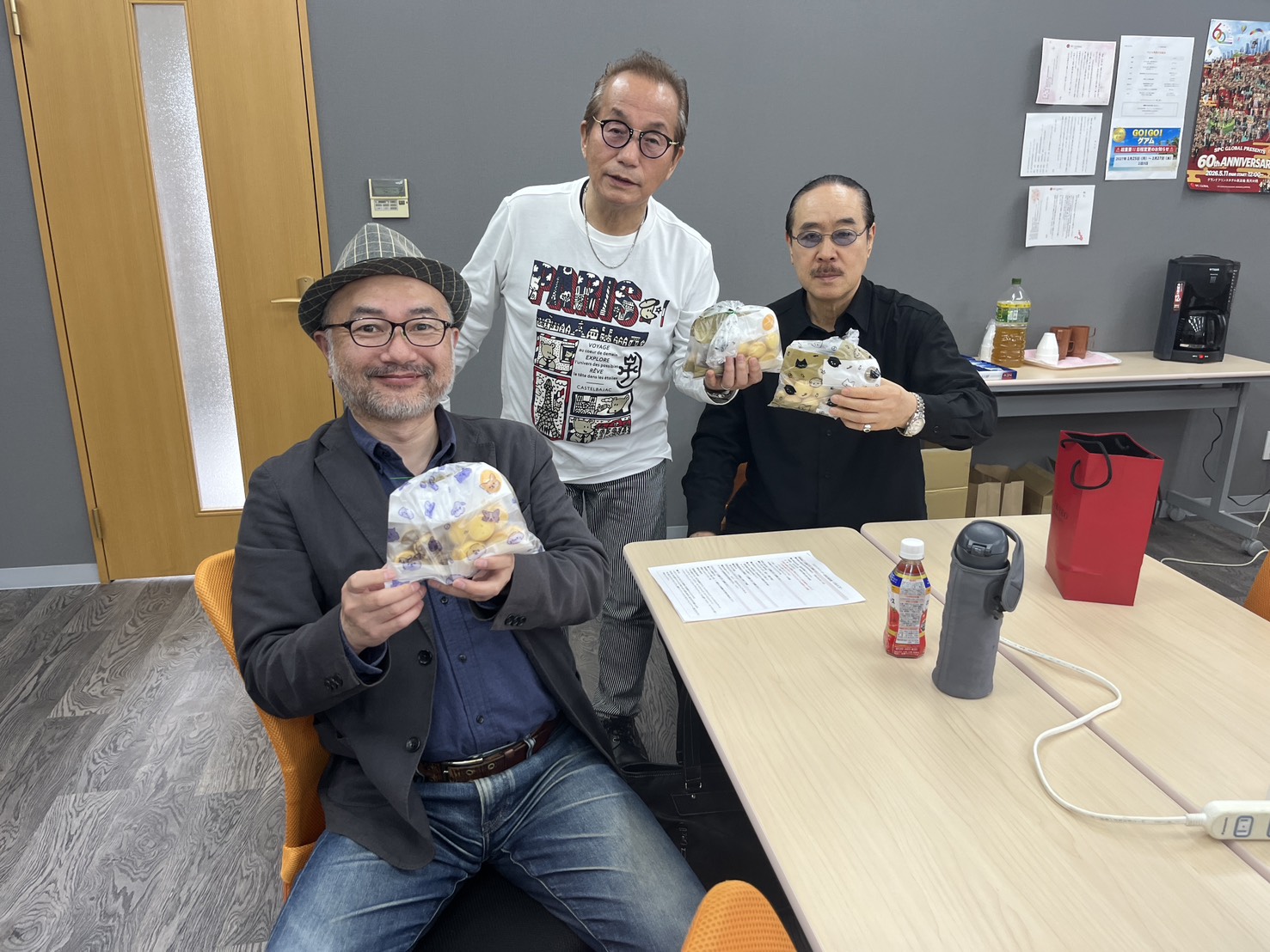Three men posing in an office, each holding wrapped snacks; table with papers and beverages in the foreground.
