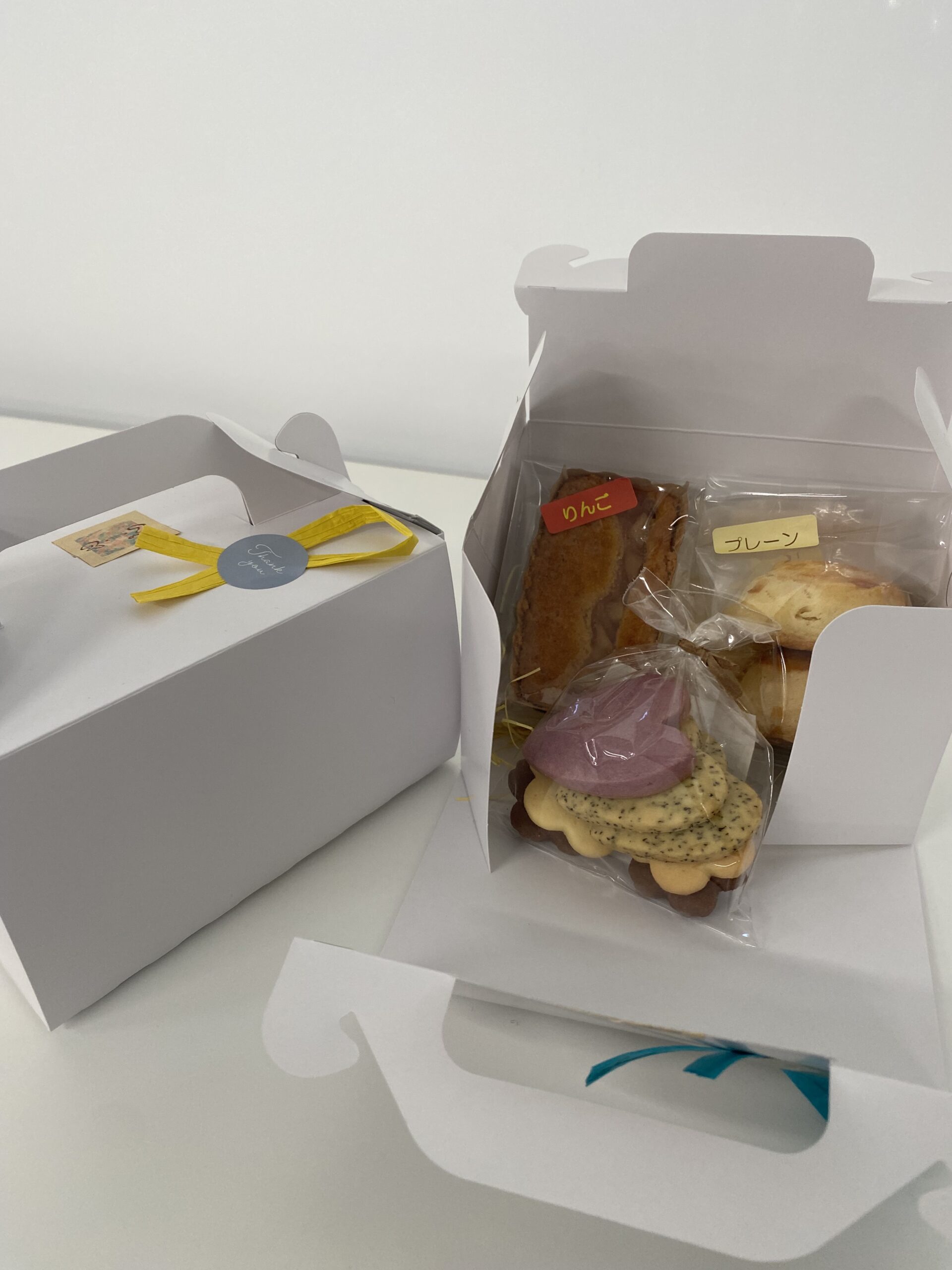 White gift boxes with a yellow ribbon next to a clear bag of cookies labeled りんご (apple) and プレーン (plain).