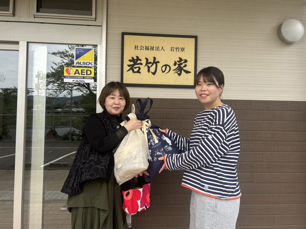 Two women exchange gift bags outside a building, standing under a gold sign with Japanese characters behind them.