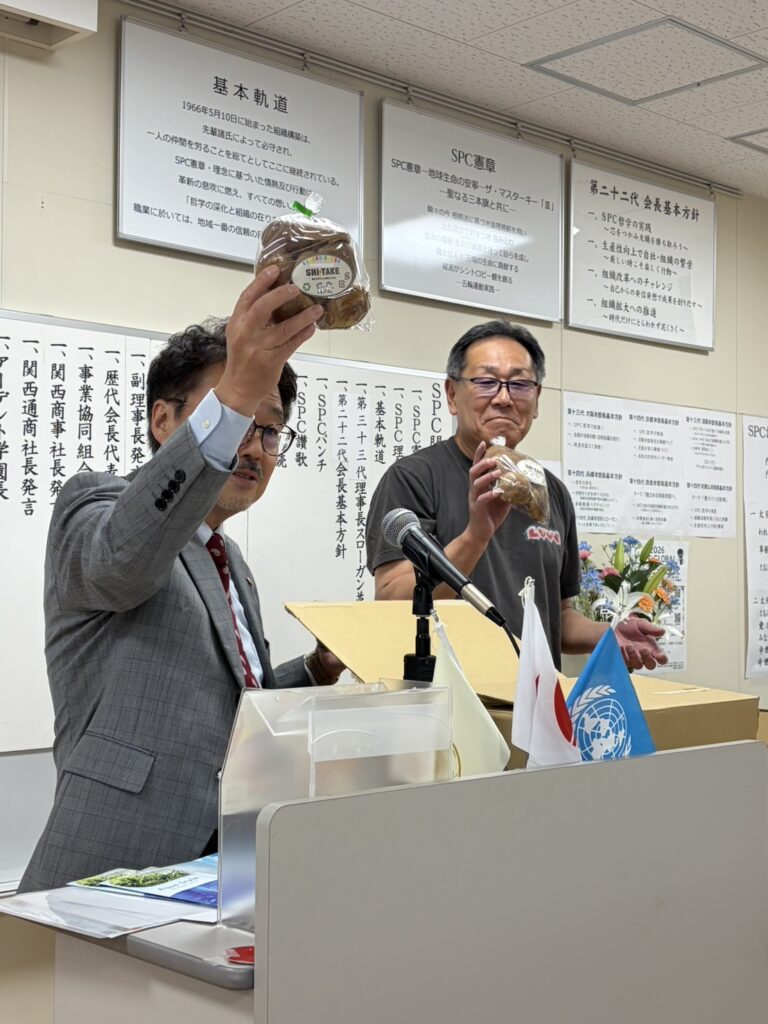 Two men at a conference desk raise packaged bread snacks labeled SHI-TAKE during a meeting attractively.