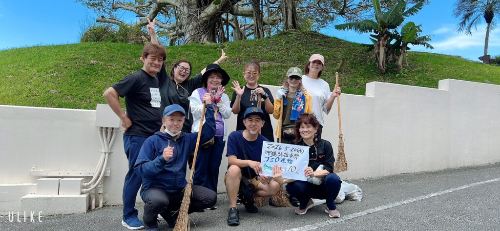 Group of ten adults posing with brooms along a sunny roadside, smiling and waving at the camera by a white wall and grassy hill nearby.