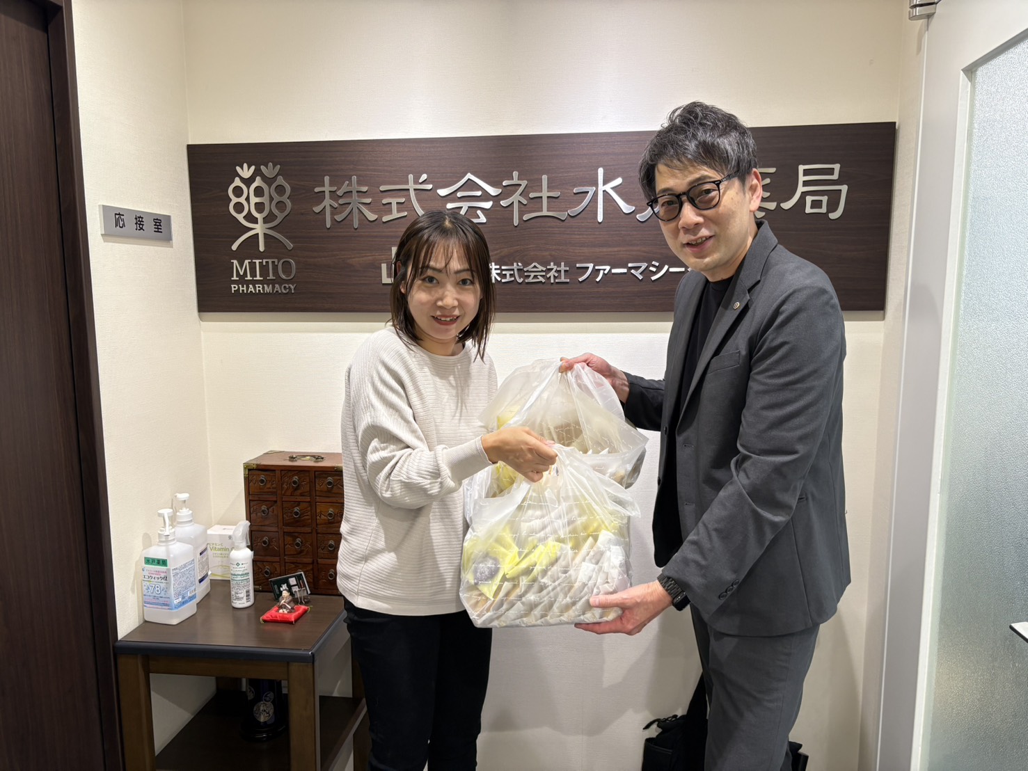 Two colleagues exchange a large transparent bag in a pharmacy lobby, standing in front of a sign reading MITO PHARMACY/水戸薬局。