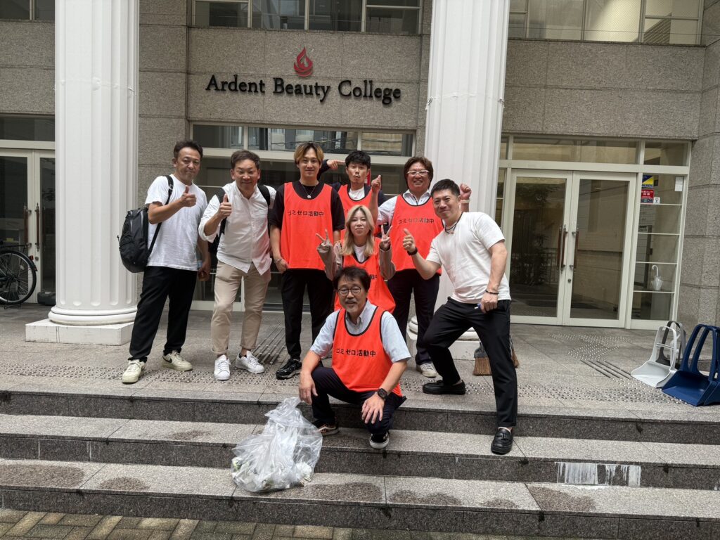 Group of volunteers in orange vests posing with thumbs up in front of Ardent Beauty College entrance; a bag of collected trash sits on the steps before them.