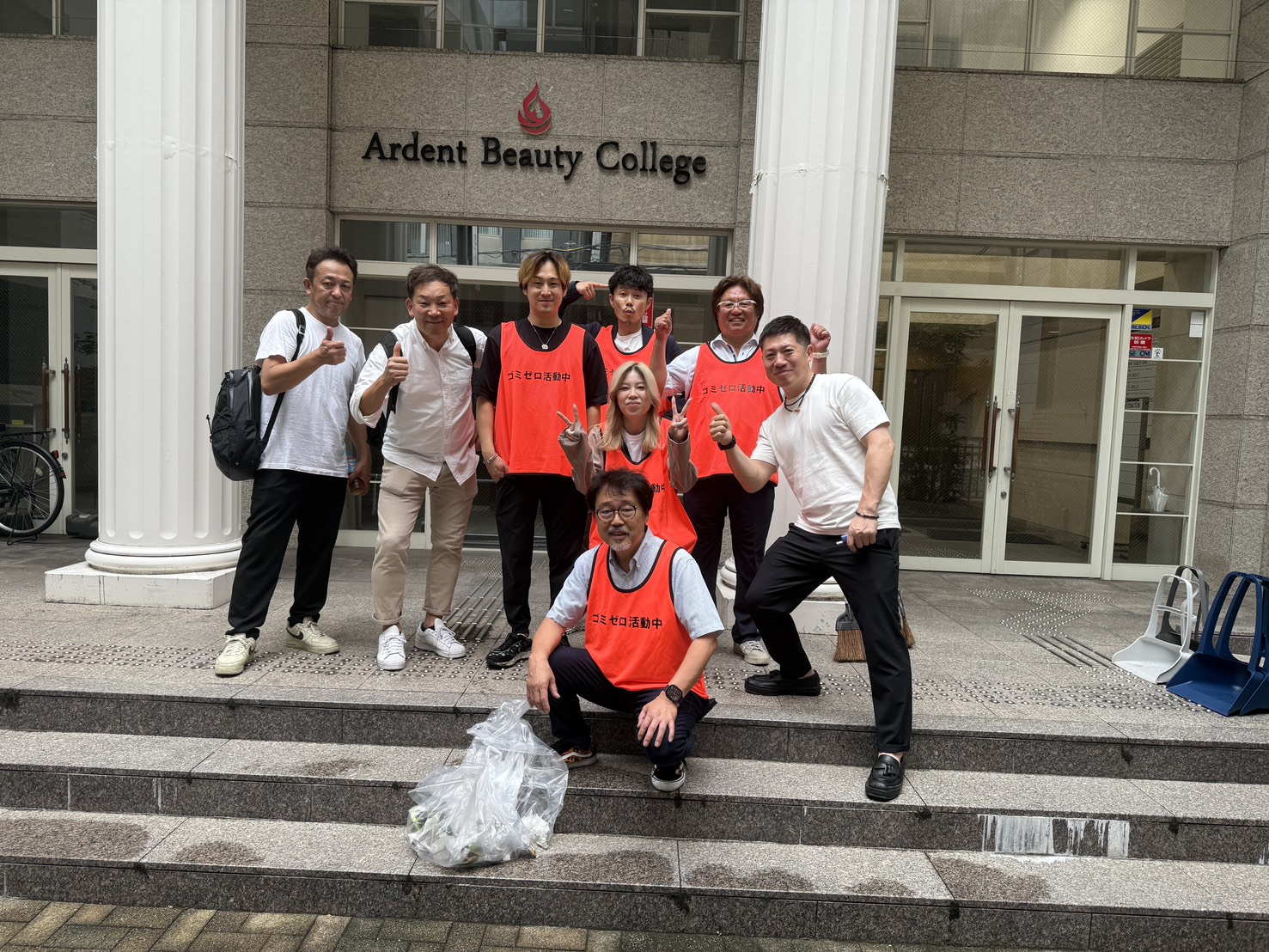Group of volunteers in orange vests posing with thumbs up in front of Ardent Beauty College entrance; a bag of collected trash sits on the steps before them.