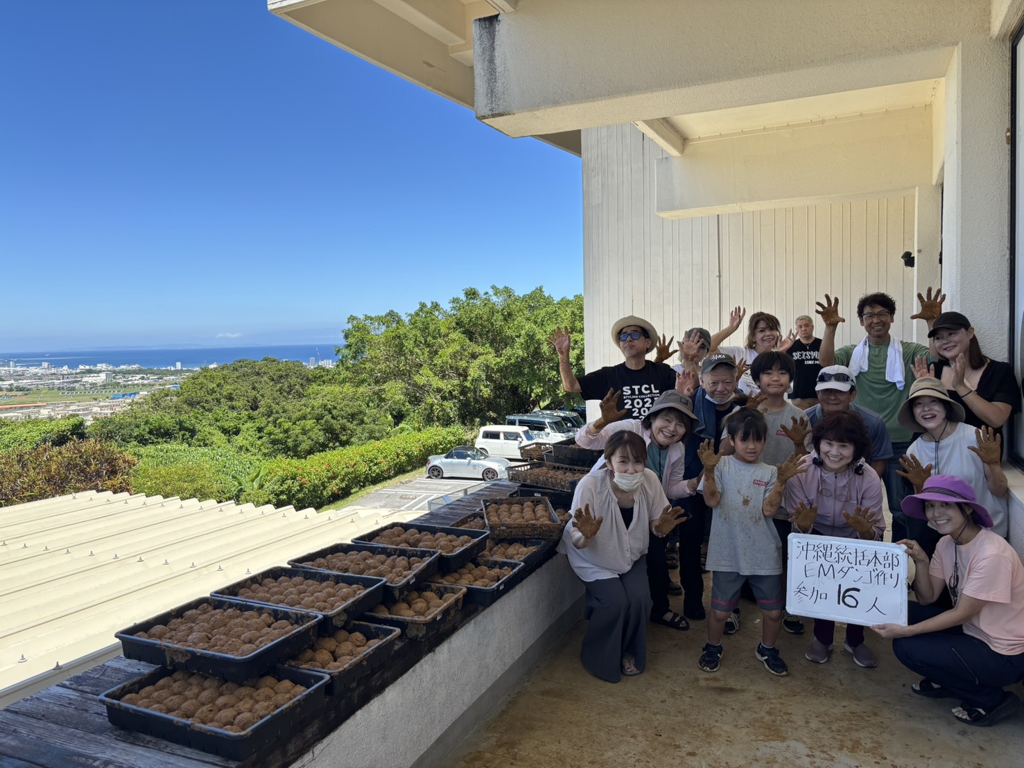 Group of 16 volunteers on a rooftop posing with trays of baked treats, waving at the camera with the ocean and city in the distance in bright sunlight.