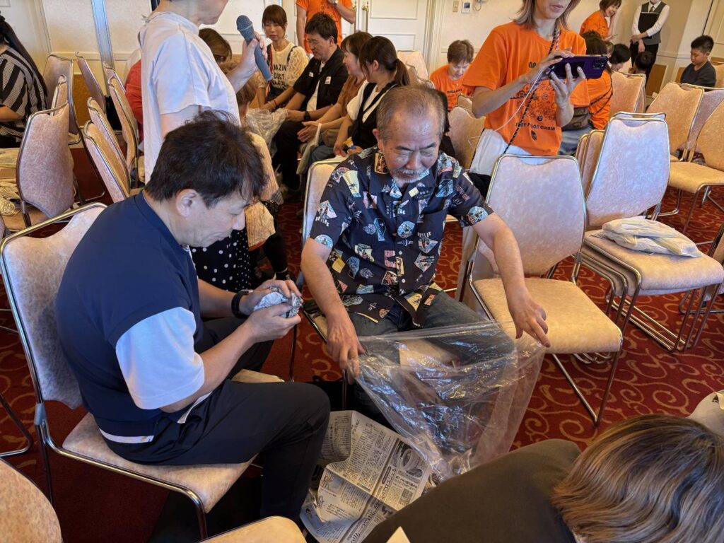 Man receiving a foot-care demo: another man handles a plastic-wrapped foot while viewers sit in armchairs at a community event, some in orange shirts in the background.