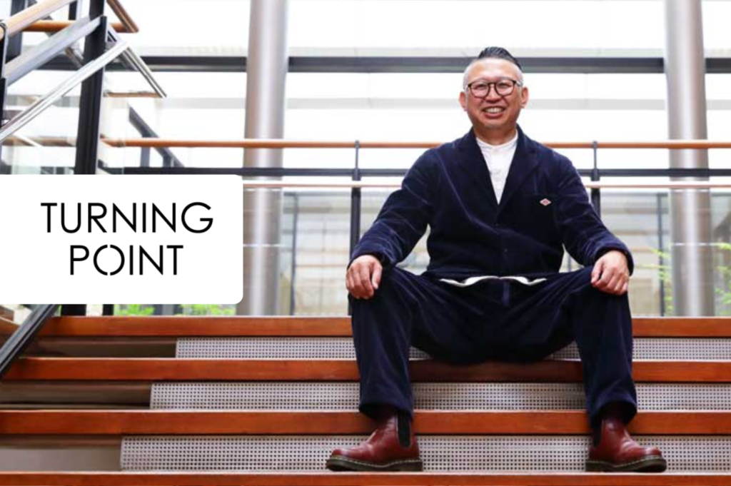 Smiling man in a navy suit sits cross‑legged on wooden stairs in a modern atrium, with a white rounded rectangle reading “TURNING POINT” on the left.