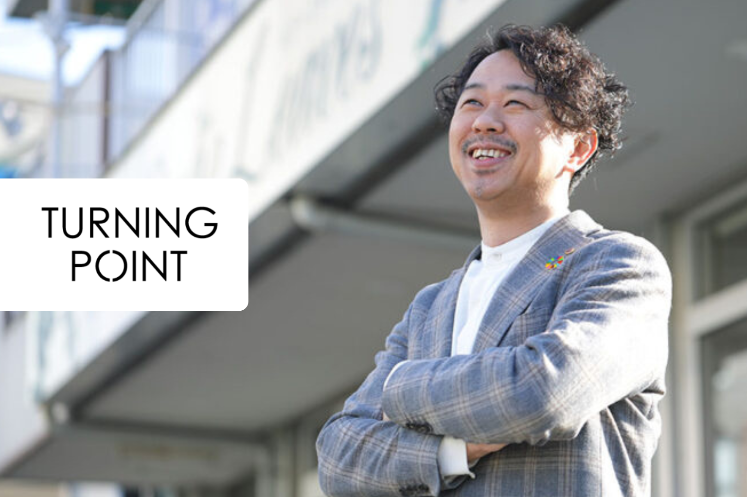 Smiling man in a gray plaid blazer stands outdoors with arms crossed; a white rounded overlay shows the text 'TURNING POINT' on the left.