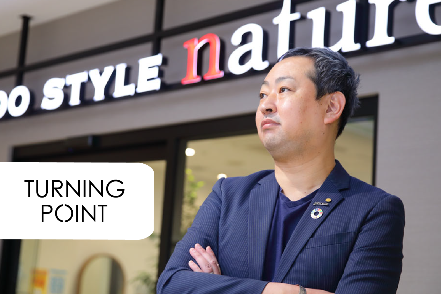 Asian man in a navy blazer stands with arms crossed outside a storefront with bold white signage; 'TURNING POINT' label overlays the image.